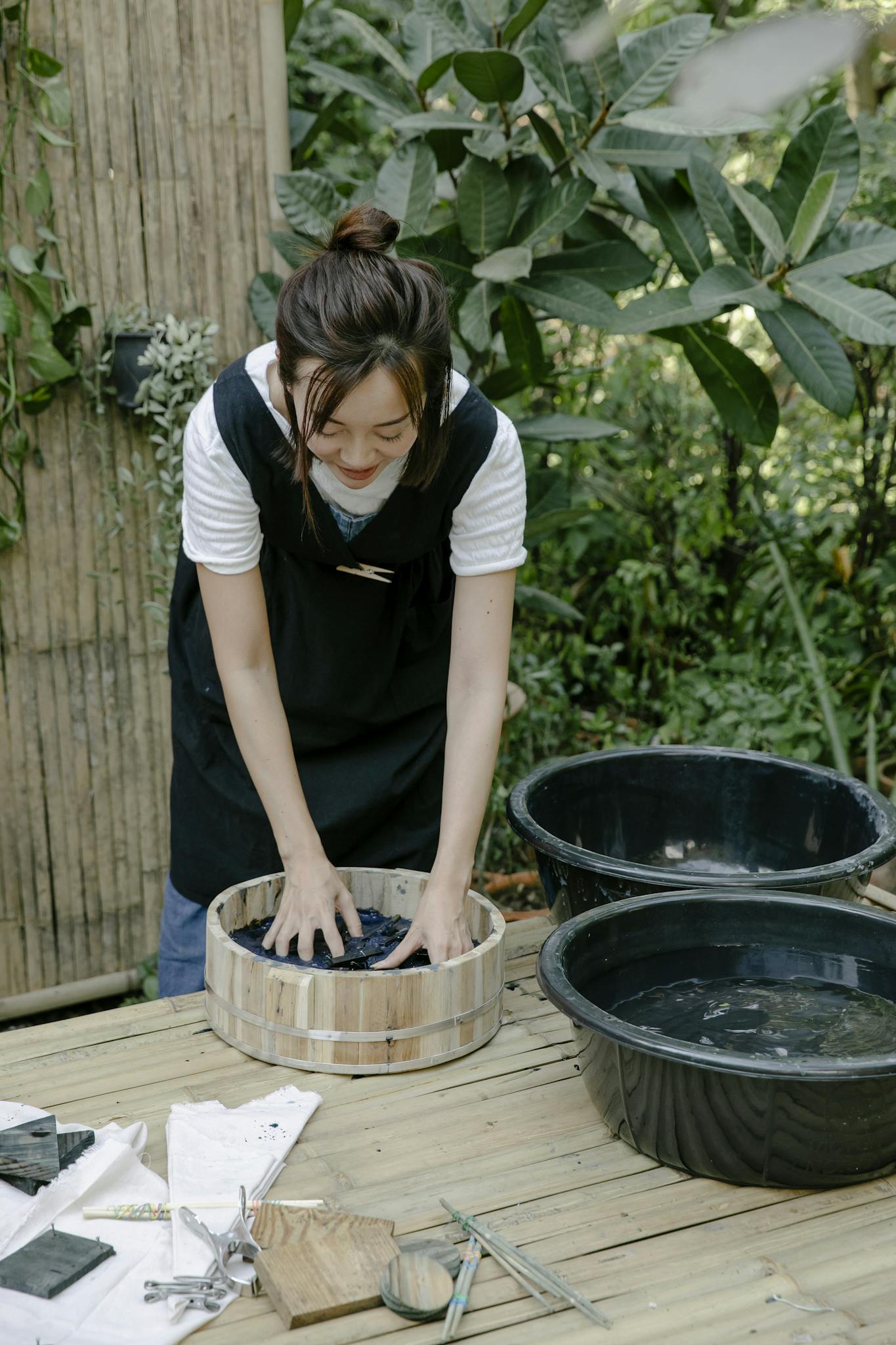 Asian woman standing outdoors tie-dyeing fabric, surrounded by greenery and natural elements.