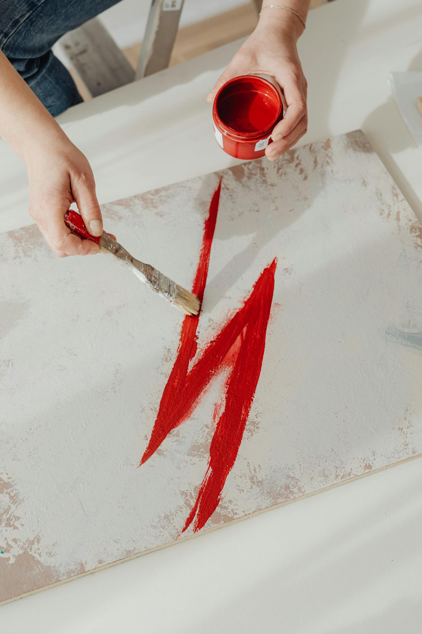 Close-up of hands painting a red brushstroke on canvas with a paintbrush and red paint can.